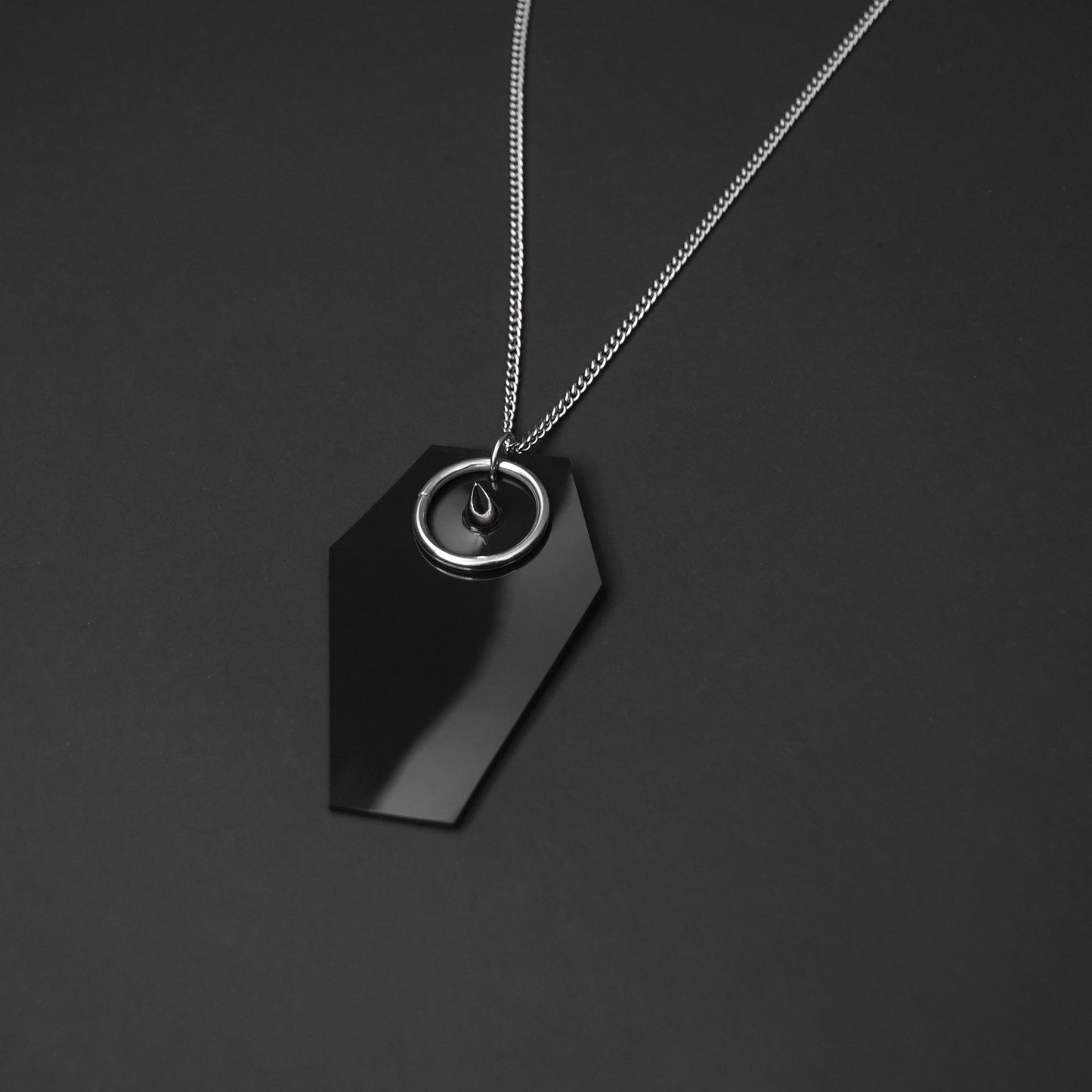 Close-up of a black PVC coffin-shaped pendant with a silver spike and O-ring, showcasing gothic-inspired details.