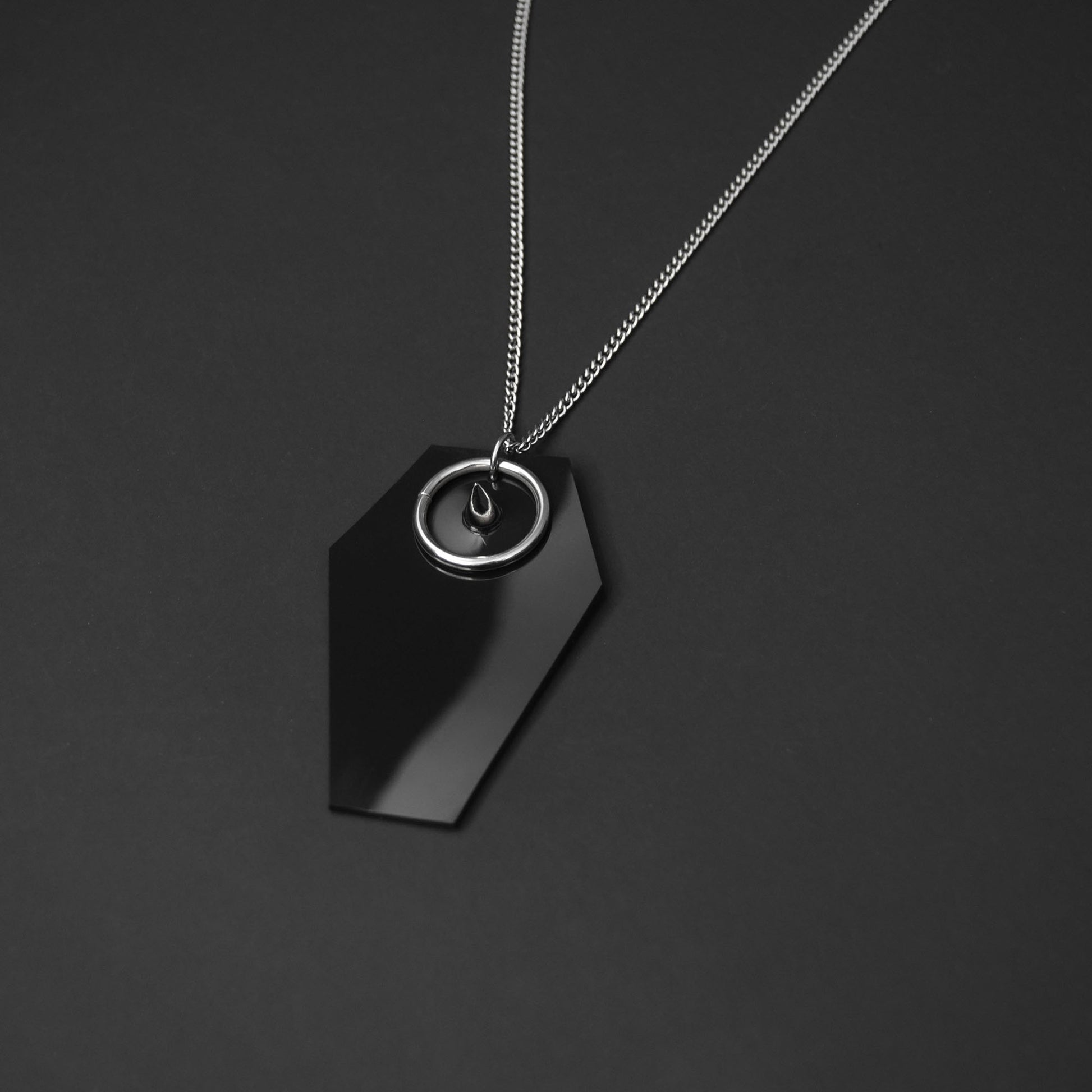 Close-up of a black PVC coffin-shaped pendant with a silver spike and O-ring, showcasing gothic-inspired details.