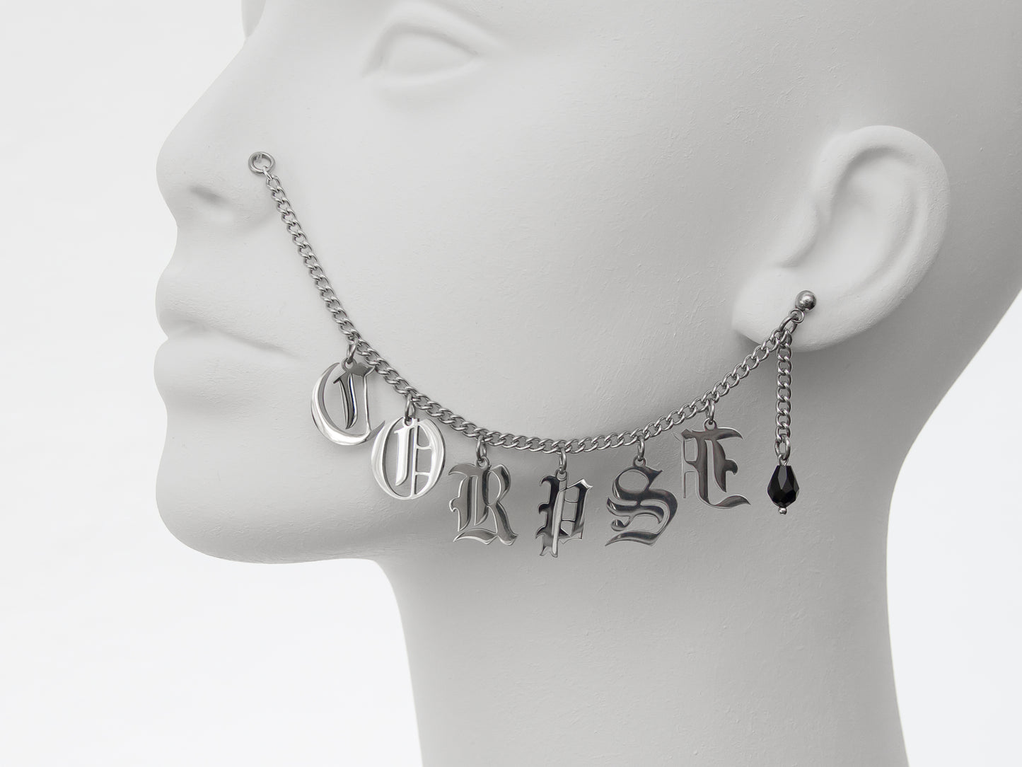 Detailed shot of silver chain nose to ear piercing with the word 'CORPSE' in Gothic-style letters, featuring a black glass teardrop earring for a dark, macabre aesthetic.