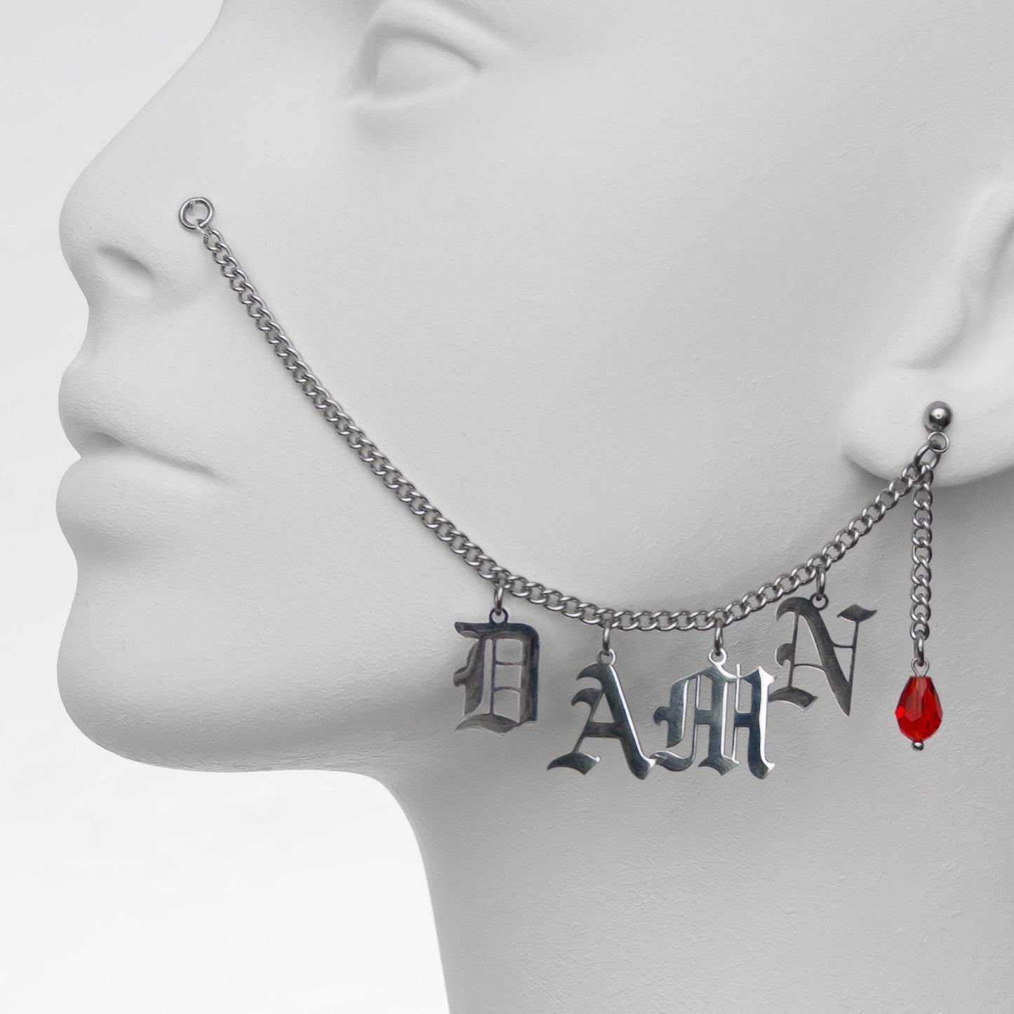 Close-up of silver chain nose to ear piercing jewellery with 'DAMN' in Gothic-style lettering, complemented by a striking red glass teardrop earring.
