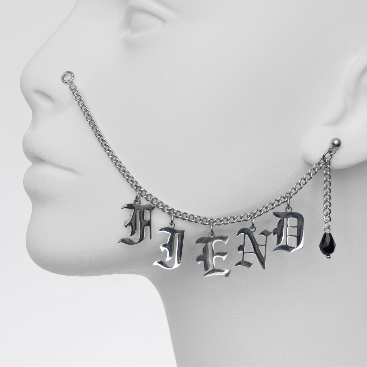 Close-up of silver chain nose to ear piercing featuring the word 'FIEND' in Gothic-style lettering, paired with a black glass teardrop earring for a rebellious, dark vibe.