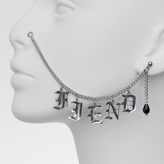 Close-up of silver chain nose to ear piercing featuring the word 'FIEND' in Gothic-style lettering, paired with a black glass teardrop earring for a rebellious, dark vibe.