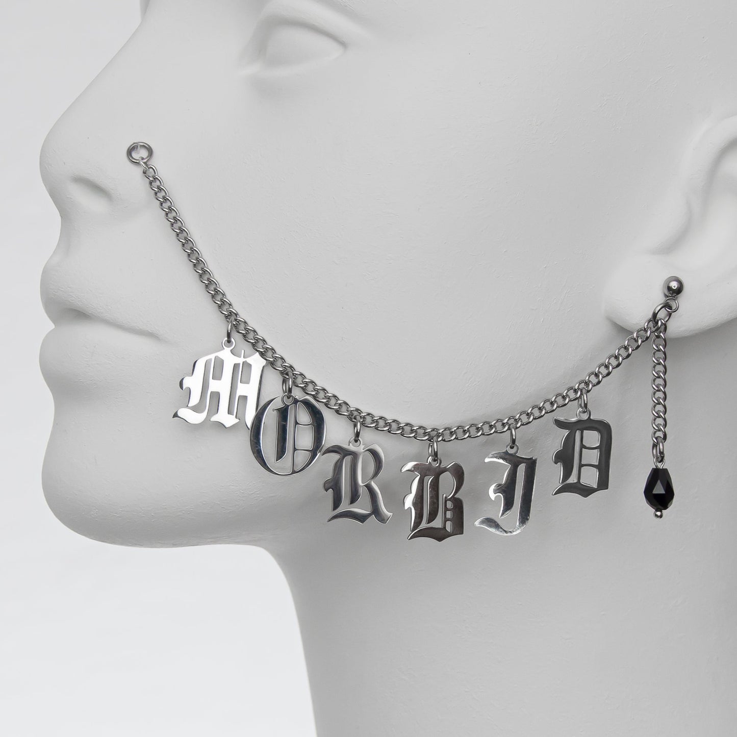 Close-up of a silver chain nose to ear piercing with 'MORBID' in Gothic-style lettering and a black glass teardrop earring, creating a dark, macabre look.