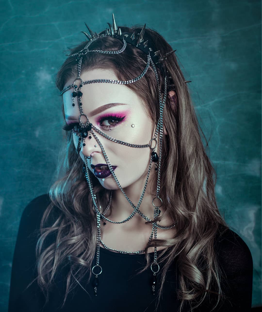 Young woman adorned with an elaborate headpiece, with metal spikes on a black PVC headband, and chains with black glass beads.