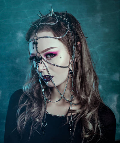 Young woman adorned with an elaborate headpiece, with metal spikes on a black PVC headband, and chains with black glass beads.