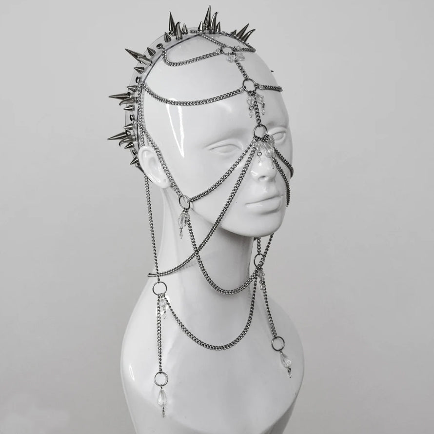 Close-up of an intricate headdress made of metal chains, clear crystals, and sharp metallic spikes, displayed on a mannequin head. The headpiece is designed to cover much of the face, set against a plain white background in a studio setting.