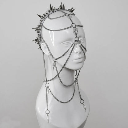 Close-up of an intricate headdress made of metal chains, clear crystals, and sharp metallic spikes, displayed on a mannequin head. The headpiece is designed to cover much of the face, set against a plain white background in a studio setting.