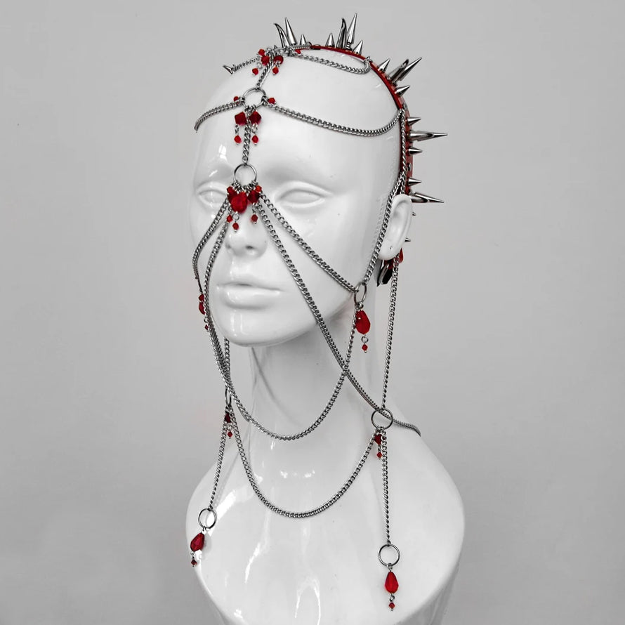 Close-up of a mannequin head adorned with an intricate headpiece made of chains, red beads, and sharp metal spikes. The background is a simple white, highlighting the gothic, edgy design of the headpiece.