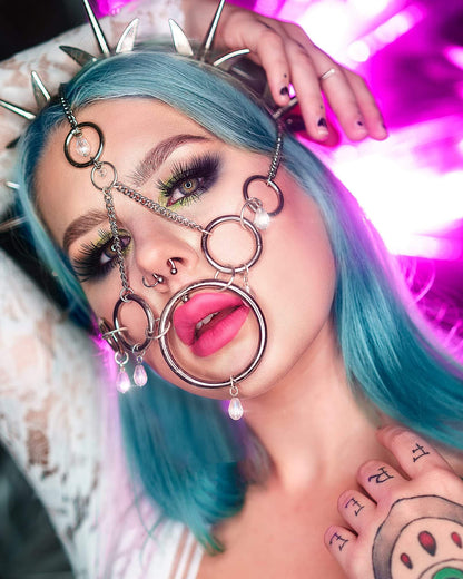 Young woman with vibrant teal hair, elaborate silver face jewellery, and a white lace top, showcasing tattoos on her hand. The background features a dreamy pink and purple light effect, emphasizing her edgy, alternative aesthetic.