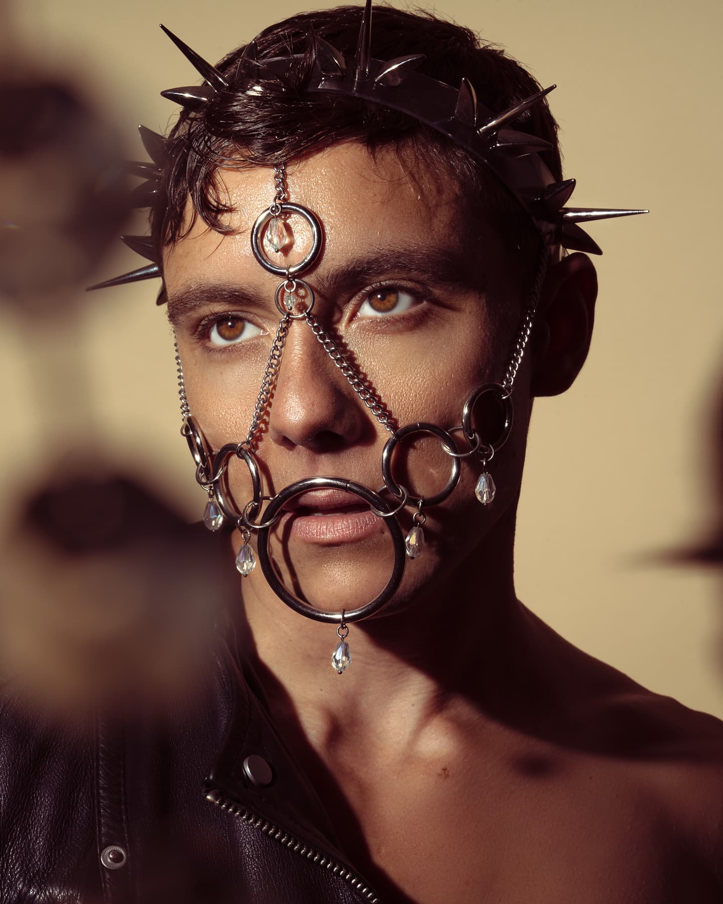 Young adult male wearing a unique metal headpiece and face chains, paired with a black leather jacket, showcasing an edgy and avant-garde aesthetic. Set against a simple background, the focus highlights intricate details of the headpiece and alternative fashion style.