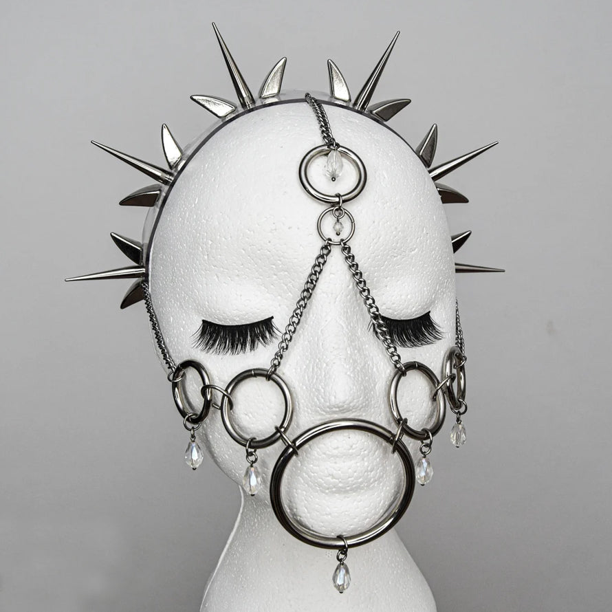 Close-up of a metal mask on a mannequin head, featuring a spiked halo, interconnected rings and chains covering the mouth and nose, and hanging crystals. The design blends gothic elegance with a rebellious aesthetic, showcased against a plain studio background.