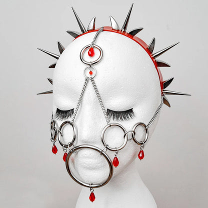 White mannequin head adorned with a striking metal and red bead headpiece, featuring a chain-link design, multiple rings, dangling beads, and a spiked headband. The intricate gothic design is highlighted against a plain background, blending dark elegance with alternative fashion.