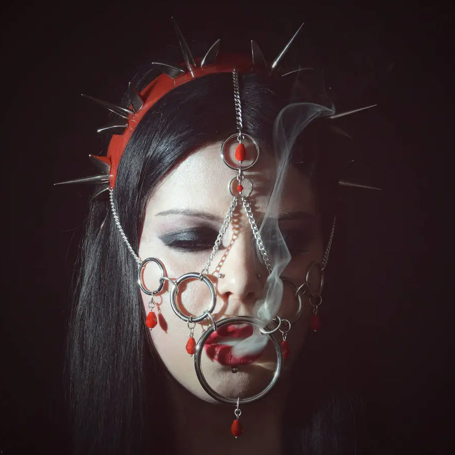 Young woman with long black hair wearing elaborate silver chains and a red spiked headband, set against a stark black background. Wisps of smoke curl from her lips, emphasizing the intricate metalwork and her gothic, mysterious aesthetic.