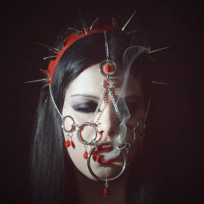 Young woman with long black hair wearing elaborate silver chains and a red spiked headband, set against a stark black background. Wisps of smoke curl from her lips, emphasizing the intricate metalwork and her gothic, mysterious aesthetic.