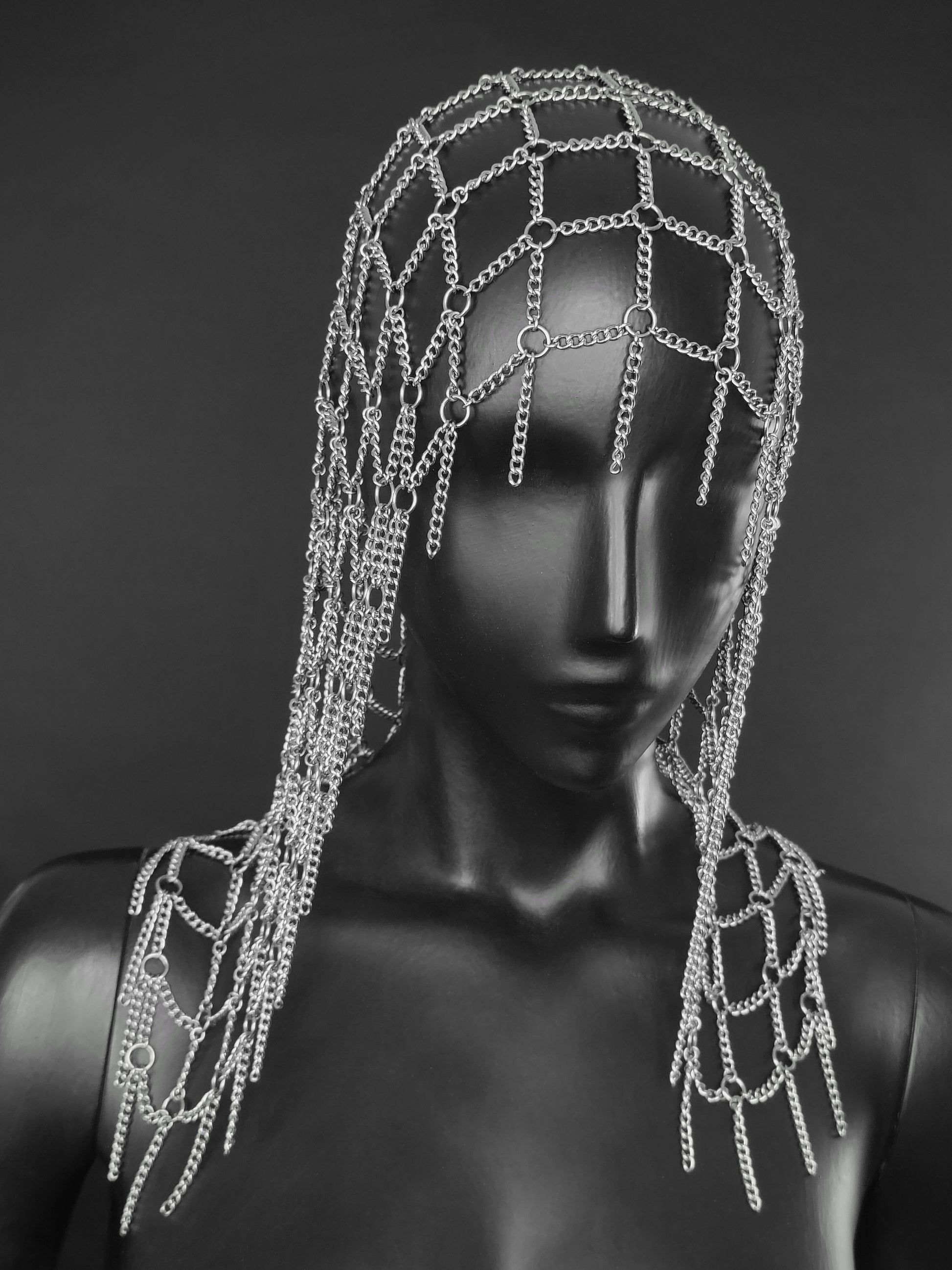 A silver chain wig like headpiece with dangling chains, displayed on a black mannequin head and shoulders against a dark gray background.