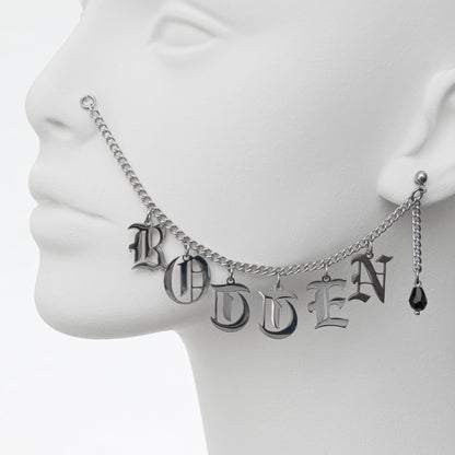 Close-up of a stylish silver chain nose to ear piercing jewellery featuring 'ROTTEN' in Gothic-style lettering and a black glass teardrop earring.