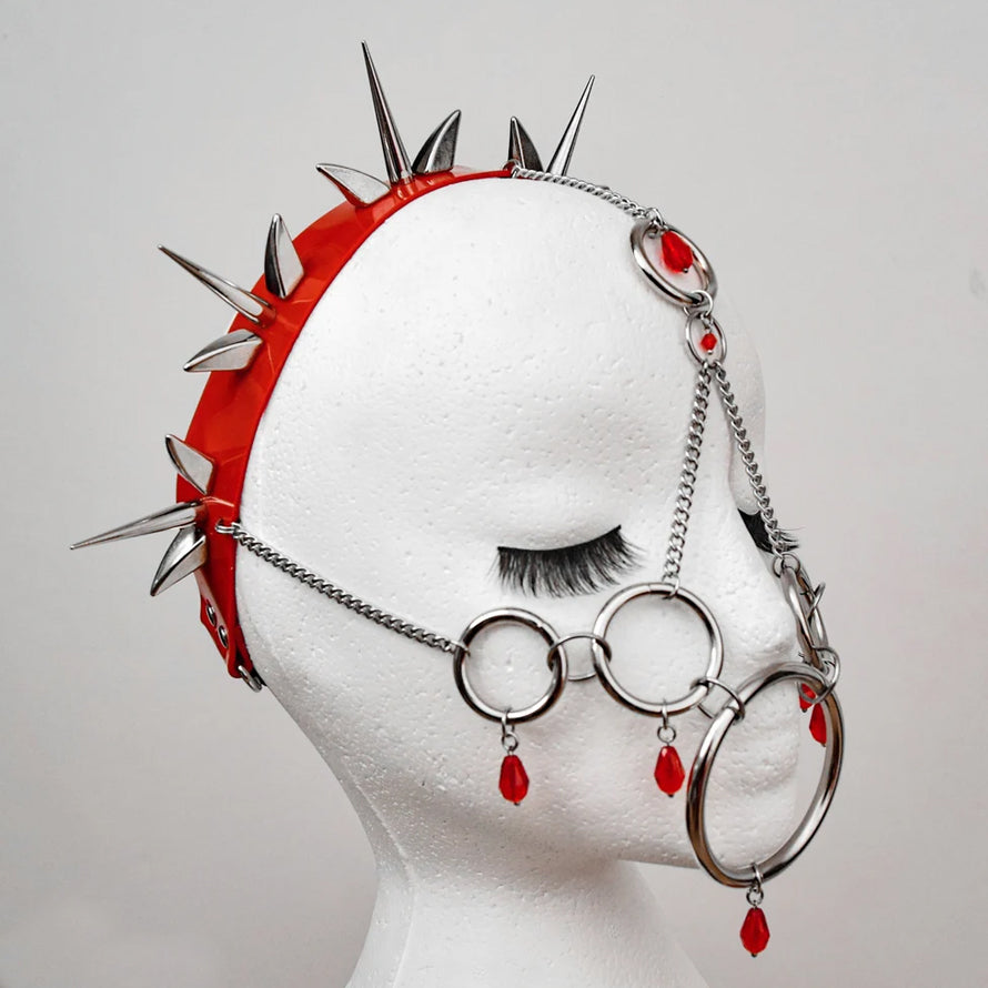Mannequin head adorned with an elaborate headpiece featuring a red PVC headband studded with sharp metal spikes, and a face mask made of interconnected metal rings and chains with red gemstone accents. The bold, gothic design is highlighted against a plain background, emphasizing its rebellious and alternative aesthetic.