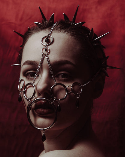 Young woman wearing a complex silver  spiked crown headpiece and face covering, set against a deep crimson background. The intricate metalwork contrasts dramatically with the backdrop, creating a bold and mysterious gothic aesthetic.