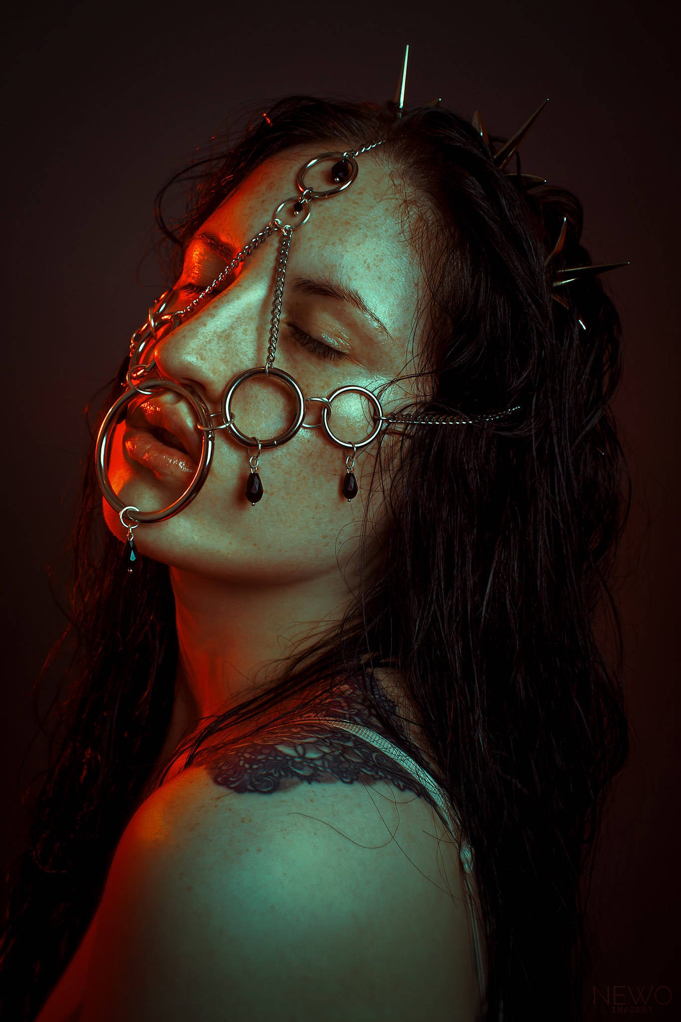 Young woman with long dark hair, wearing an intricate metal spiked crown headpiece and facial harness. Set against a dark studio background, her serene expression highlights the elaborate design and gothic-inspired aesthetic.