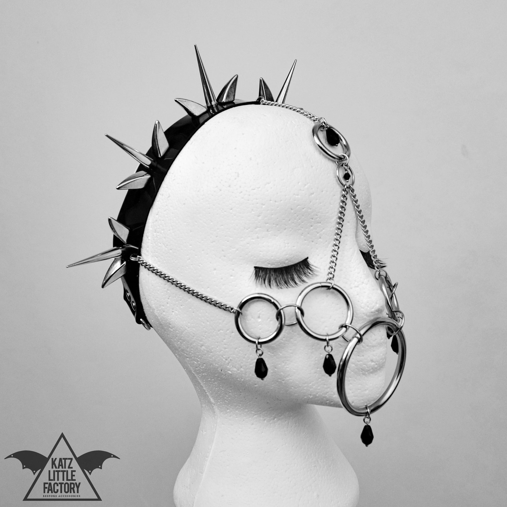 Mannequin head wearing a striking black PVC headband with sharp metallic spikes and delicate chains forming interconnected rings that partially obscure the face. The bold gothic and edgy design is highlighted against a neutral background, emphasizing its alternative aesthetic.