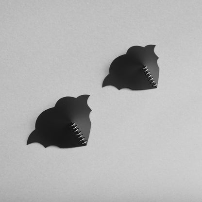 A pair of gothic black heart PVC breast petals with symmetrical bat wings and a central row of jump rings. at an angle.