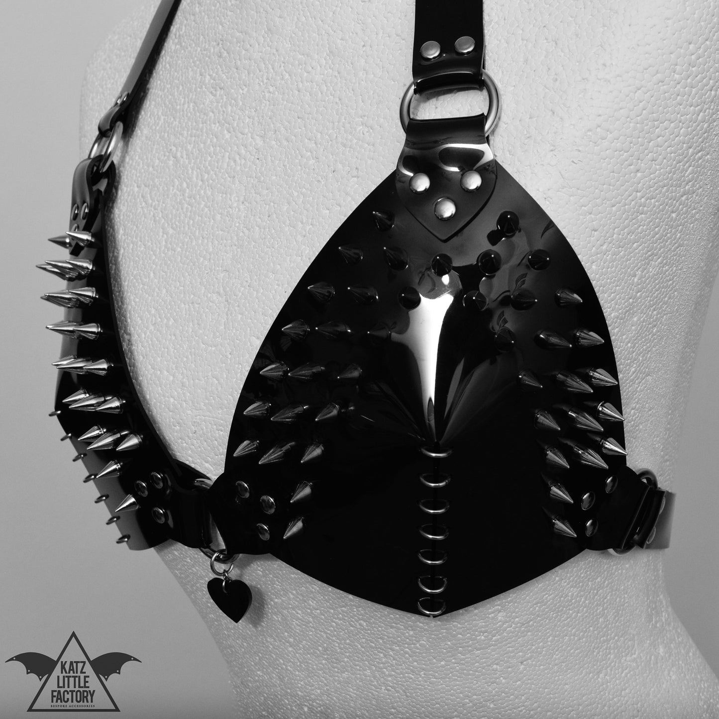 This photo features a fashion accessory bra. The piece is made of glossy black PVC with spikes protruding from it, featuring a heart shaped pendant.